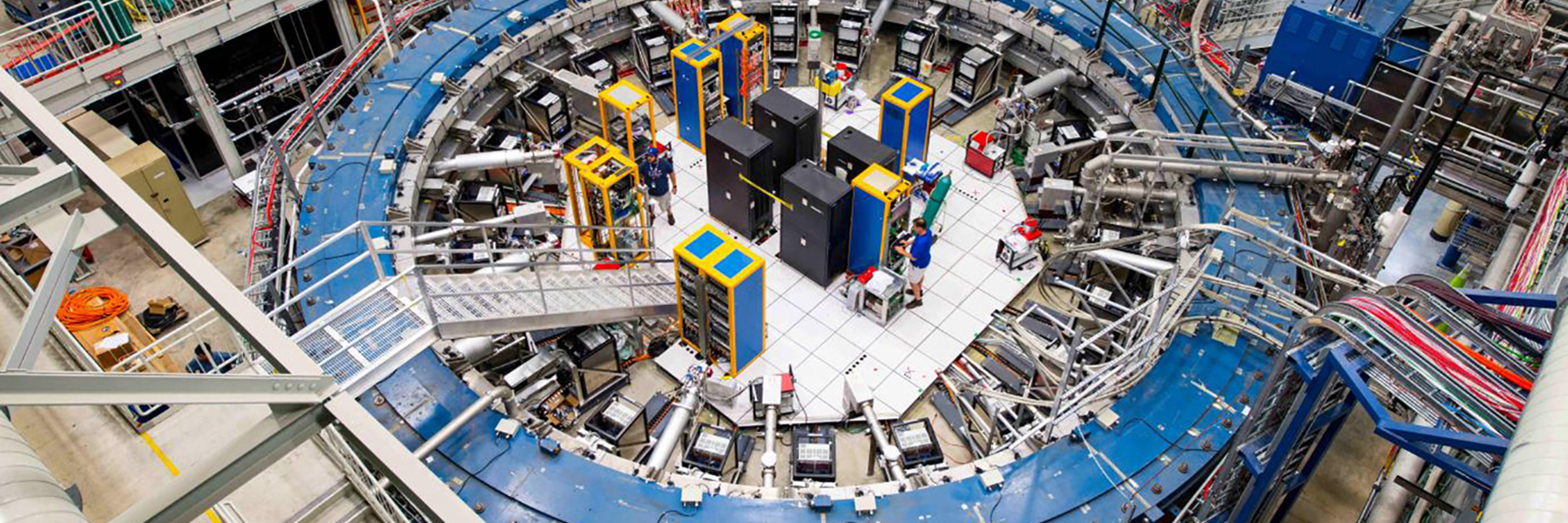 Image of the Muon G2 experiment hall at Fermi National Accelerator Laboratory taken by Reidar Hahn. 
                    The image shows a large technology room from above. Pictured is the muon accelerator, which appears as a huge blue tubular circle. 
                    The muon ring is connected to many computers and different types of equipment that are located in the center of the ring. 
                    There appear to be stairs that go up and over the tube from one side to the other for access for the scientists. (photo by Reidar Hahn Fermilab)