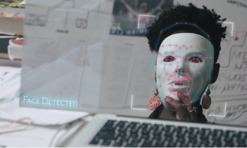 This is an image to promote the film Coded Bias. Included is a picture of a black female who is sitting in front of a computer screen. Over her face, she holds a white mask which has red dots and lines to indicate facial recognition. 